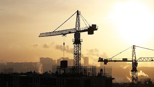 Foto, zwei Kräne sowie ein Rohbau als schwarze Silhouetten im Gegenlicht der Sonne. Am Horizont rauchende Fabrikschornsteine.