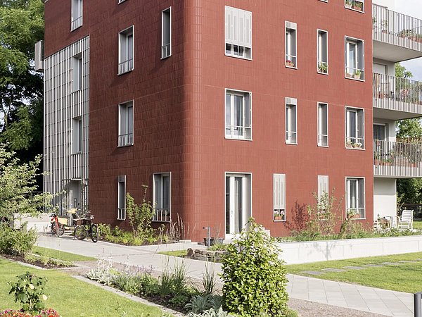 Dreieckiges, rotes Wohngebäude mit mehreren Fenstern und Balkonen, umgeben von einem Garten mit Pflanzen und Wegen.