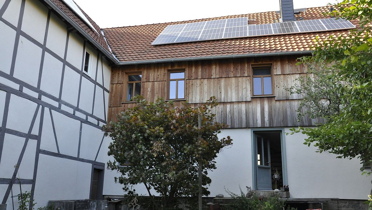 Foto, Rückansicht eines Hauses mit Holzverkleidung und Solarzellen auf dem Dach, umgeben von Bäumen und Garten