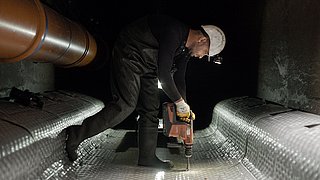 Foto, ein Arbeiter mit Helm steht in einem Tunnel und bohrt in den Boden.