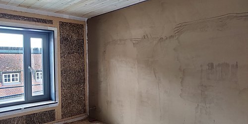 Foto, Innenraum mit einer aus Stroh gefertigten Wand mit Fenster und einer mit Lehm verputzten Wand. 