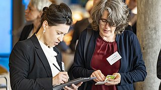 Foto, zwei Frauen unterhalten sich auf einer Fachveranstaltung, die eine macht sich Notizen, die andere hält eine Karte und ein Namensschild.