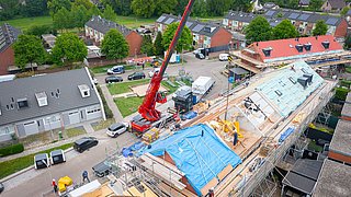 Foto, eine Baustelle mit einem roten Kran, Holzkonstruktionen und einer blauen Abdeckung auf einem Hausdach in einer Wohnsiedlung.