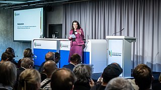 Foto, Frau in pinkem Kostüm hält Notizen und spricht vor Publikum in einem Konferenzraum mit grauen Vorhängen und Präsentationsbildschirm.