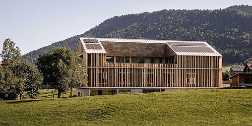 Foto, modernes, langgestrecktes Holzgebäude mit großen Fenstern und Solarpaneelen auf dem Dach vor bewaldetem Hügel