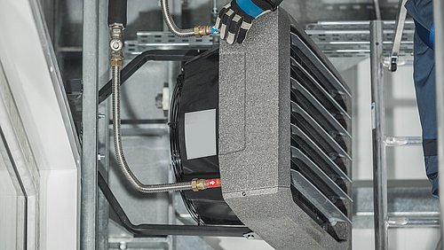 Foto, Ein Techniker mit Handschuhen installiert ein schwarzes Heizaggregat, das mit Metallrohren und Armaturen befestigt ist, an einer Metallstruktur.
