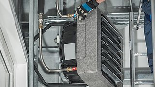 Foto, Hand mit Arbeitshandschuh hält graues, rechteckiges Lüftungsgerät mit Metallrohren in technischer Umgebung