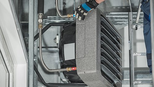 Foto, Hand mit Arbeitshandschuh hält graues, rechteckiges Lüftungsgerät mit Metallrohren in technischer Umgebung