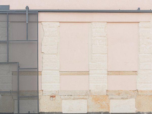 Foto, Fassade mit drei zugemauerten Fenstern, davor gepflasterter Boden und ein Beet mit kleinen Pflanzen und gelben Blumen.