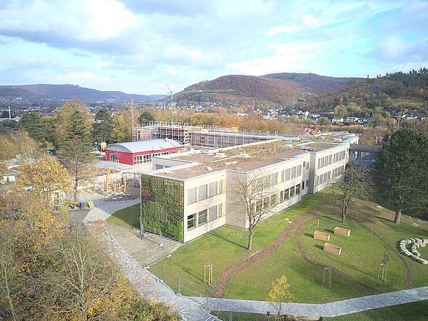 Luftaufnahme eines Schulcampus mit modernem, begrüntem Flachdachgebäude, umgeben von Bäumen, Wegen und Sitzgelegenheiten, im Hintergrund Hügel und Wohnhäuser.