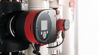 Foto, Nahaufnahme eines runden, schwarzen und roten elektronischen Geräts mit Display und Tasten an einer Rohrleitung.