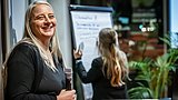 Foto, Frau mit langem, blondem Haar hält Mikrofon, im Hintergrund eine weitere Frau, die an einem Flipchart schreibt.