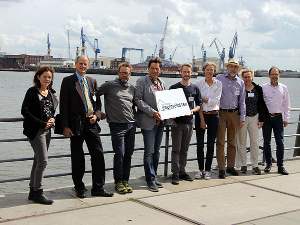 Foto, Gruppenbild mehrerer Personen, die nebeneinander an einem Geländer lehnen. Dahinter befindet sich ein Fluss und der Hamburger Hafen.