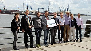 Foto, Gruppenbild mehrerer Personen, die nebeneinander an einem Geländer lehnen. Dahinter befindet sich ein Fluss und der Hamburger Hafen.