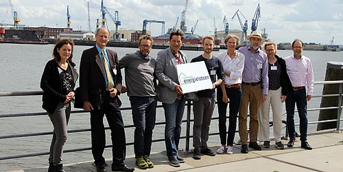 Foto, Gruppenbild mehrerer Personen, die nebeneinander an einem Geländer lehnen. Dahinter befindet sich ein Fluss und der Hamburger Hafen.