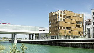 Foto, Holzbau mit mehreren Balkonen am Ufer eines Kanals, daneben moderne weiße Brücke