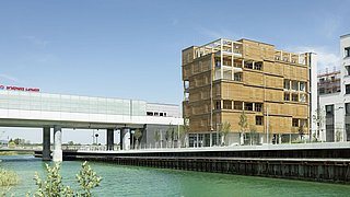 Foto, Holzbau mit mehreren Balkonen am Ufer eines Kanals, daneben moderne weiße Brücke