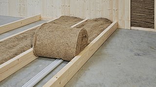 Foto, Rolle aus brauner Schafwolle zur Bodenisolierung auf Betonboden mit Holzkonstruktion und Rohrleitungen