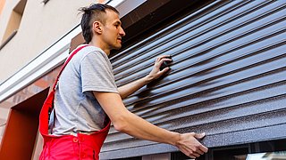 Foto, Handwerker in roter Latzhose und grauem T-Shirt montiert eine Außenjalousie an einem Gebäude