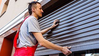 Foto, Handwerker in roter Latzhose und grauem T-Shirt montiert eine Außenjalousie an einem Gebäude