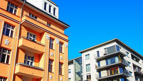 Foto, Zwei Wohngebäude, eines orange mit Balkonen und Verzierungen, das andere weiß mit Glasaufzug und Balkonen