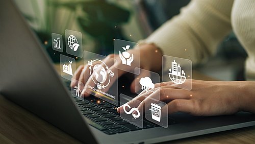 Foto, Nahaufnahme einer Person, die an einem Schreibtisch sitzend an einem Laptop arbeitet; über dem Laptop schweben digital hinzugefügte Symbole zum Thema Lebenszyklus.