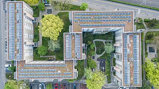 Foto, Luftaufnahme eines Gebäudekomplexes mit begrünten Dächern, Solaranlagen, Parkplätzen und einem Innenhof mit Bäumen und Wegen.