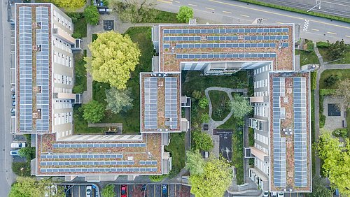 Foto, Luftaufnahme eines Gebäudekomplexes mit begrünten Dächern, Solaranlagen, Parkplätzen und einem Innenhof mit Bäumen und Wegen.