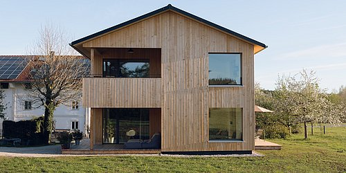 Modernes zweistöckiges Holzhaus mit großen Fenstern und Balkon, umgeben von Wiese und Bäumen, daneben ein weißes Haus mit Solarpanelen.