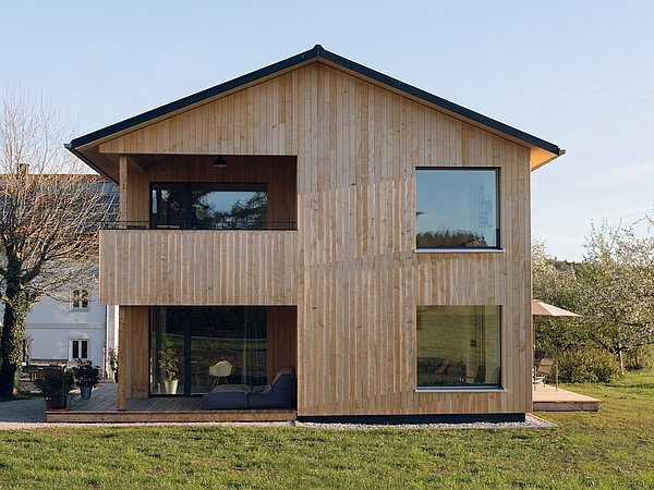 Modernes zweistöckiges Holzhaus mit großen Fenstern und Balkon, umgeben von Wiese und Bäumen, daneben ein weißes Haus mit Solarpanelen.