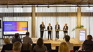 Foto, eine Podiumsdiskussion mit vier Rednern an Stehtischen in einem hellen Raum mit großen Fenstern und einem gesprächsfreudigen Publikum.