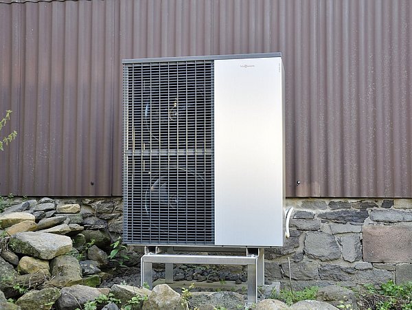 Foto, Wärmepumpe, freistehend auf Metallgestell vor einer Steinmauer und brauner Fassade