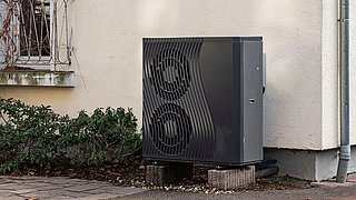 Foto, außenstehende Wärmepumpe mit zwei großen Lüftern an einer Hauswand neben einem Fenster und Pflanzen