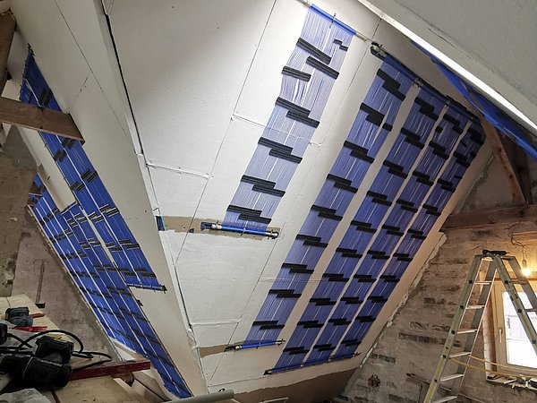 Foto, Dachgeschoss mit installierten blauen Deckenflächenheizungen unter Schräge, Bauleiterleiter und Baumaterialien sichtbar