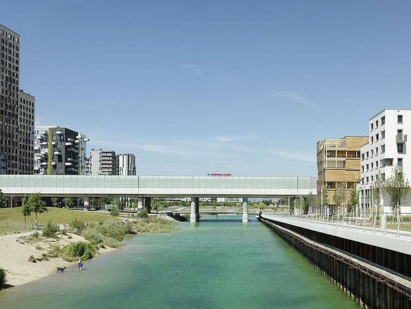 Foto, Blick auf einen Kanal mit moderner Fußgängerbrücke und Wohngebäuden links und rechts unter blauem Himmel