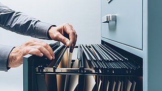 Seitliche Nahaufnahme der offenen Schublade eines Aktenschrankes. Zwei Hände greifen in die Schublade und durchsuchen die Akten.