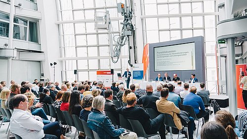Foto, Menschen verfolgen einen Vortrag an einem Messestand.