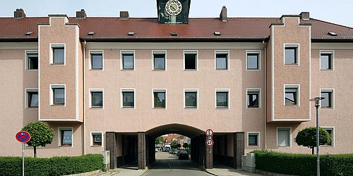 Foto, Frontalansicht auf dreistöckiges Mehrfamilienhaus mit Haus-Unterführung im Erdgeschoss und Glockenturm auf dem Dach.