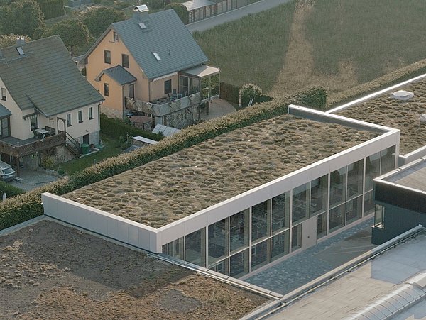 Foto, Luftaufnahme eines modernen Flachdachgebäudes mit begrüntem Dach neben traditionellen Wohnhäusern in einer ländlichen Umgebung.