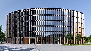 Foto, Außenansicht des Rathauses in Freiburg, einem mehrstöckigen, runden Gebäudes mit einer von Holzpanele und Photovoltaikmodulen durchbrochenen Glasfassade.