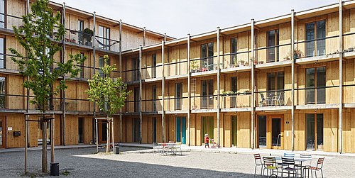 Foto, Innenhof eines dreistöckigen Wohngebäudes mit Holzfassade und Balkonen, Tischen und Stühlen auf Kiesboden