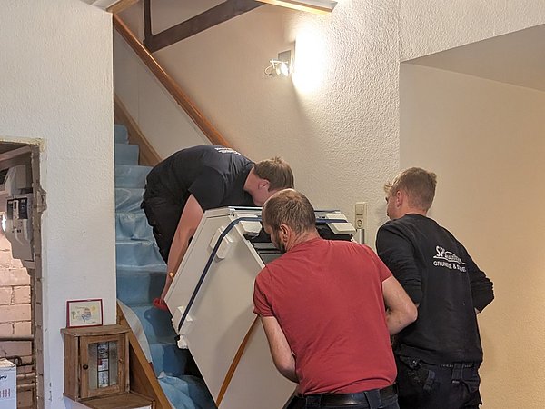 Foto, Drei Männer tragen gemeinsam eine große weiße Wärmepumpe eine Treppe hinauf