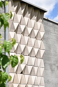 Foto, Fassade mit dreidimensionalen, dreieckigen Mustern neben einer glatten Wand, links grüne Blätter im Vordergrund.