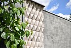 Foto, Fassade mit dreidimensionalen, dreieckigen Mustern neben einer glatten Wand, links grüne Blätter im Vordergrund.