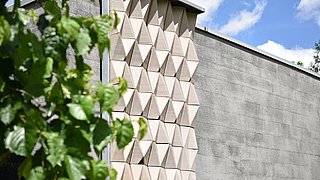 Foto, Fassade mit dreidimensionalen, dreieckigen Mustern neben einer glatten Wand, links grüne Blätter im Vordergrund.