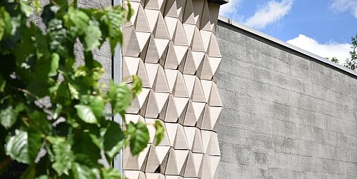 Foto, Fassade mit dreidimensionalen, dreieckigen Mustern neben einer glatten Wand, links grüne Blätter im Vordergrund.