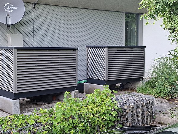 Foto, Zwei große, rechteckige Wärmepumpengeräte auf Betonplatten vor einem Haus mit Fenster und Satellitenschüssel