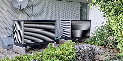 Foto, Zwei große, rechteckige Wärmepumpengeräte auf Betonplatten vor einem Haus mit Fenster und Satellitenschüssel