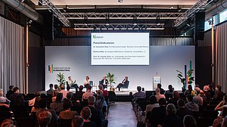 Foto, Blick aus dem Publikum auf eine Bühne auf der eine Podiumsdiskussion mit mehreren Teilnehmenden stattfindet.