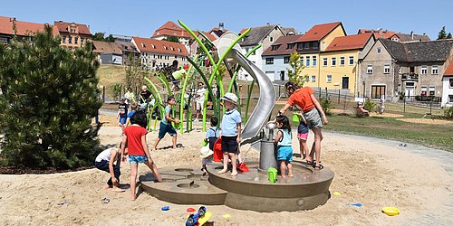Foto, Kinder spielen an einem Wasserspielplatz mit Rutsche und Sand in einem Wohngebiet mit bunten Häusern im Hintergrund